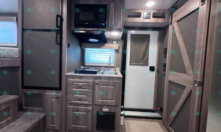 Interior of a modern RV kitchen: fridge, microwave, stove with vent hood, sink, and wood cabinets on a compact counter. A doorway and stairs are to the left.