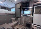 Interior of a compact RV: bed on the left, steps up, tall fridge, microwave, small kitchen with sink and cabinets.