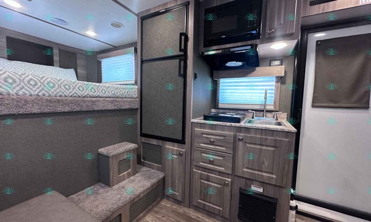 Interior of a compact RV: bed on the left, steps up, tall fridge, microwave, small kitchen with sink and cabinets.