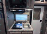 Compact RV kitchen with microwave above a stove and small sink on a speckled countertop, wood-tone cabinets all around