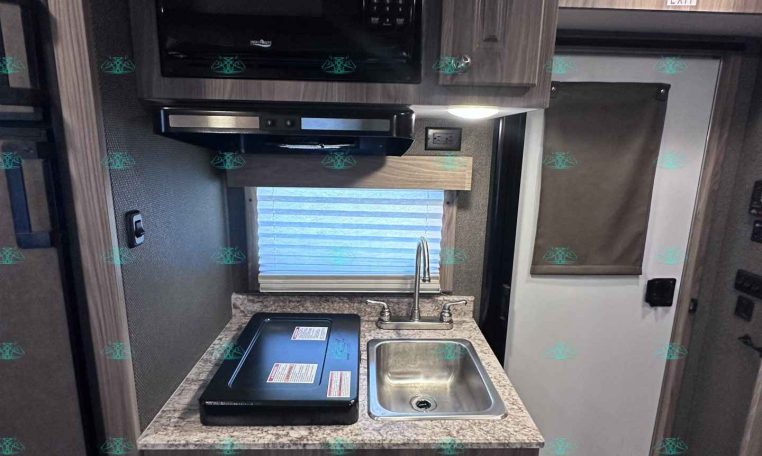 Compact RV kitchen with microwave above a stove and small sink on a speckled countertop, wood-tone cabinets all around