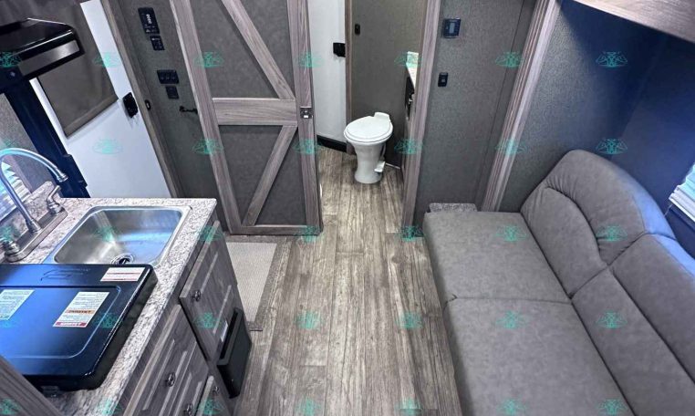 Interior of a compact camper trailer: kitchen counter with sink on left, sofa along right, wooden cabinets, and a small bathroom at the back.