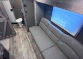 Interior of a travel trailer with a gray sectional sofa, upper wood cabinets, and a small bathroom with a toilet visible at the rear.