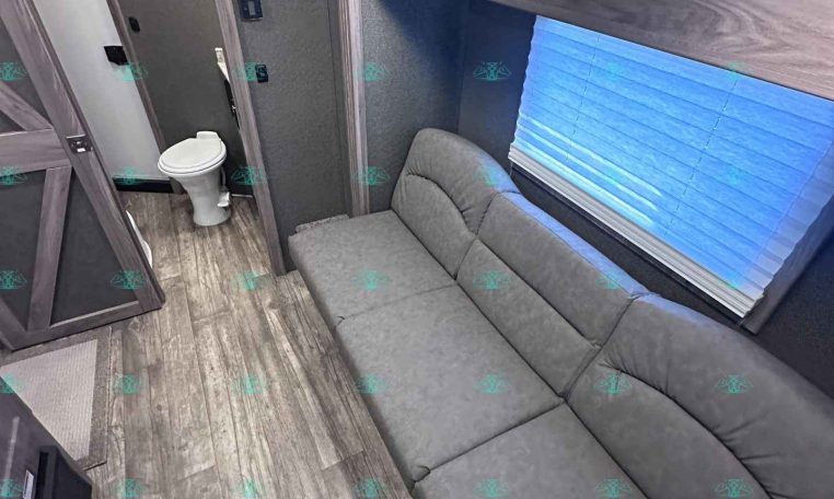Interior of a travel trailer with a gray sectional sofa, upper wood cabinets, and a small bathroom with a toilet visible at the rear.