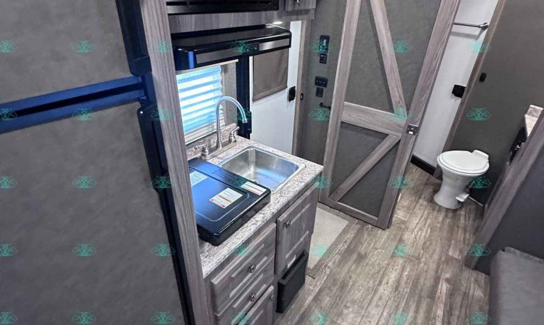 RV kitchenette with microwave above a counter and sink, wood-tone cabinets, and a sliding barn-style door leading to a bathroom area