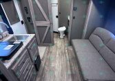 Interior of a modern travel trailer: kitchen on left with sink, stove, and cabinets; living area with a gray sofa on the right; bathroom door and toilet straight ahead.