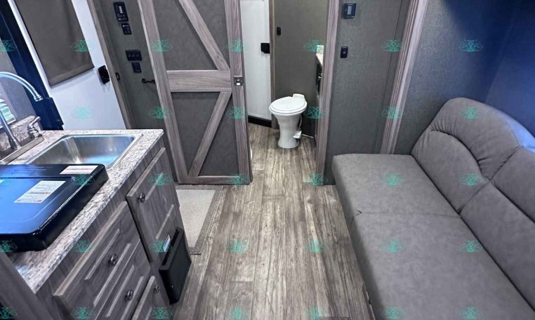 Interior of a modern travel trailer: kitchen on left with sink, stove, and cabinets; living area with a gray sofa on the right; bathroom door and toilet straight ahead.