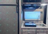 Interior of a compact RV kitchen: a tall stainless fridge on the left, microwave above a countertop with a sink and faucet, and wooden cabinets and drawers surrounding the workspace.