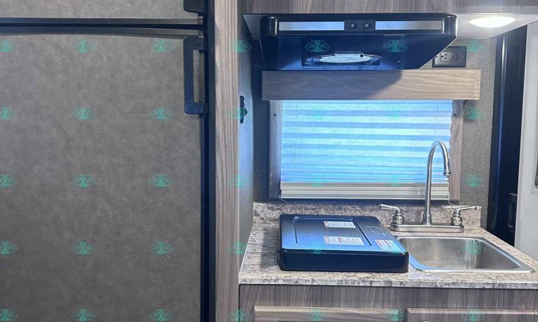 Interior of a compact RV kitchen: a tall stainless fridge on the left, microwave above a countertop with a sink and faucet, and wooden cabinets and drawers surrounding the workspace.