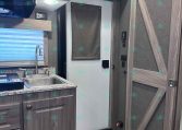 Compact RV kitchen with wood cabinets, stainless sink, and faucet; gray mini-fridge in center, window with blind, and ceiling light above.