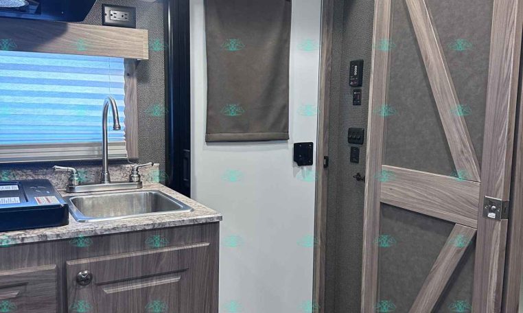 Compact RV kitchen with wood cabinets, stainless sink, and faucet; gray mini-fridge in center, window with blind, and ceiling light above.