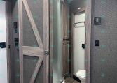 RV bathroom with a glass-curtain shower enclosure and wood-paneled sliding doors, visible toilet, and towel bar.
