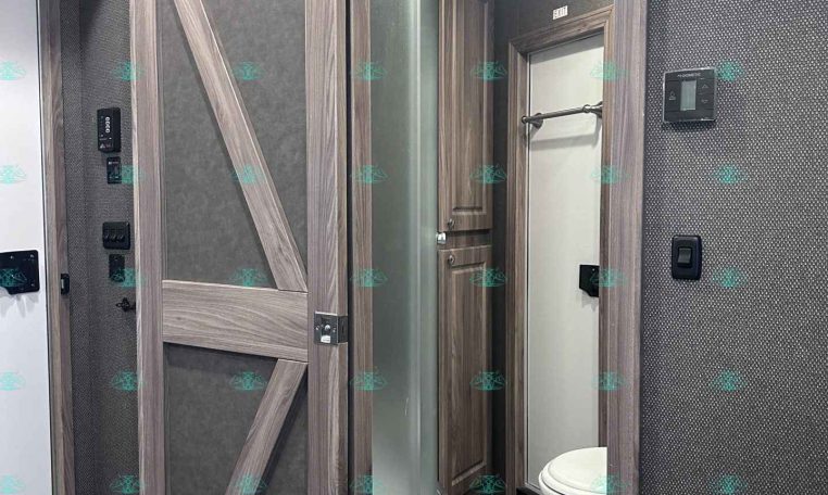 RV bathroom with a glass-curtain shower enclosure and wood-paneled sliding doors, visible toilet, and towel bar.