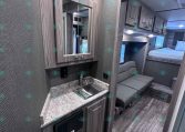 Interior of a modern RV/kitchen area with grey wood cabinets, a stainless sink, and marble-like countertop, reflected in a wall mirror next to a seating nook and bunk beds in the background