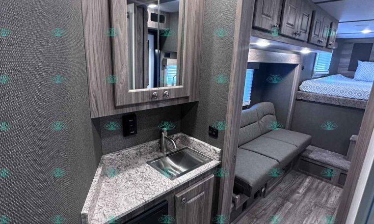 Interior of a modern RV/kitchen area with grey wood cabinets, a stainless sink, and marble-like countertop, reflected in a wall mirror next to a seating nook and bunk beds in the background
