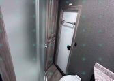 Small bathroom inside a trailer with a curved glass shower enclosure, wooden cabinet, and toilet nearby.