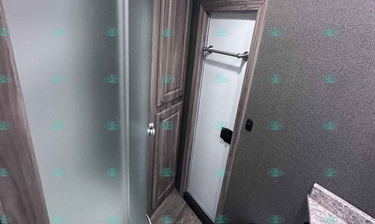 Small bathroom inside a trailer with a curved glass shower enclosure, wooden cabinet, and toilet nearby.