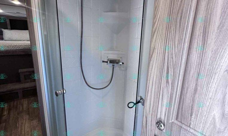 RV shower stall with curved glass doors, white tiled walls, and a chrome handheld shower on a flexible hose near a woodgrain cabinet.