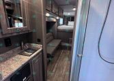 Interior of a compact travel trailer showing a kitchenette with sink, granite countertop, and upper cabinets, leading to a living area with sofa and a bed in the back.