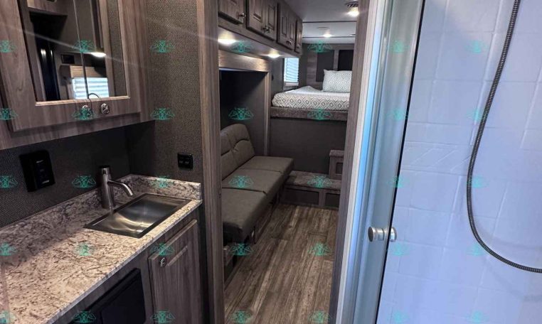 Interior of a compact travel trailer showing a kitchenette with sink, granite countertop, and upper cabinets, leading to a living area with sofa and a bed in the back.