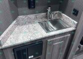 Compact trailer kitchen: stainless sink with curved faucet, marblelike countertop, gray wood cabinets, and a small wall control panel behind the sink.