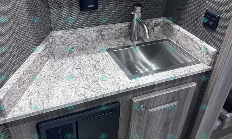 Compact trailer kitchen: stainless sink with curved faucet, marblelike countertop, gray wood cabinets, and a small wall control panel behind the sink.