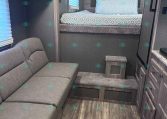 RV bedroom with raised bed, teal-patterned bedding, and a gray upholstered headboard area with storage cabinets above.
