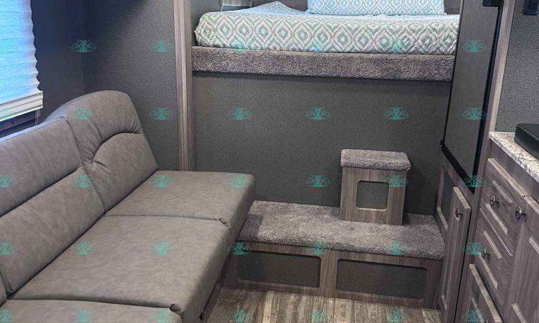 RV bedroom with raised bed, teal-patterned bedding, and a gray upholstered headboard area with storage cabinets above.