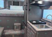 Compact RV interior with a bed at the rear, patterned bedding, and stair-step access to the platform area with storage below.
