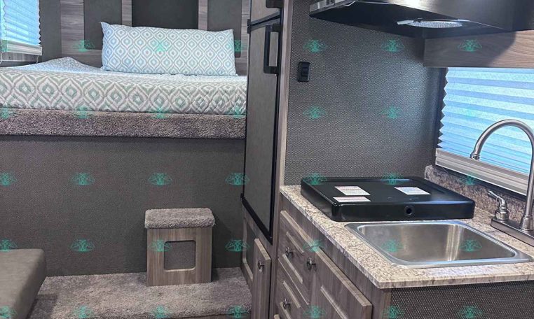Compact RV interior with a bed at the rear, patterned bedding, and stair-step access to the platform area with storage below.