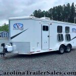 White horse trailer with Charger by Lakota logo parked on gravel lot, trees in background.”] ,