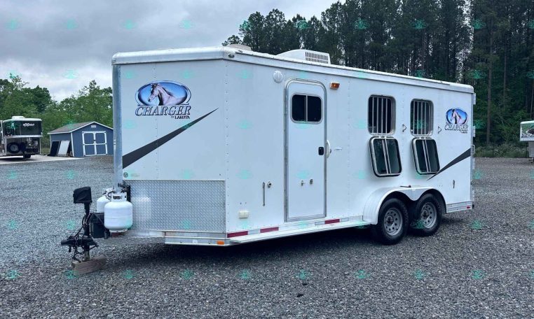 White horse trailer with Charger by Lakota logo parked on gravel lot, trees in background.”] ,
