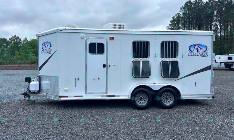 White horse trailer with two axles and barred windows parked on a gravel lot under a cloudy sky, Charger by Lakota logo on the side.