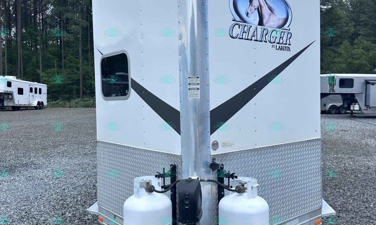 Front view of a white horse trailer with Charger by Lakota logo and a horse head graphic, two propane tanks at the hitch on a gravel lot.