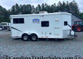 White horse trailer with Charger by Lakota logo sits on a gravel lot, with a red tractor on the right and trees in the background.
