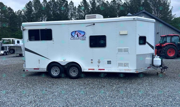 White horse trailer with Charger by Lakota logo sits on a gravel lot, with a red tractor on the right and trees in the background.