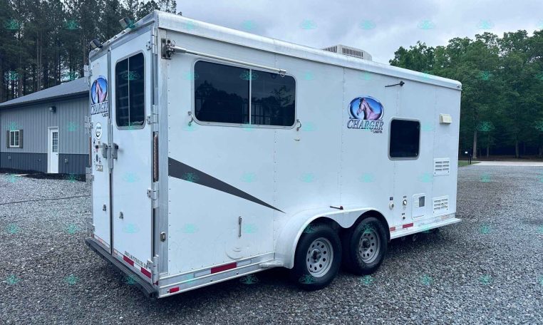 White horse trailer with Charger logo parked on a gravel lot with trees in the background.
