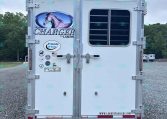 Rear of a white Lakota Charger horse trailer with a large horse logo and various stickers, parked on gravel with trees in the background.