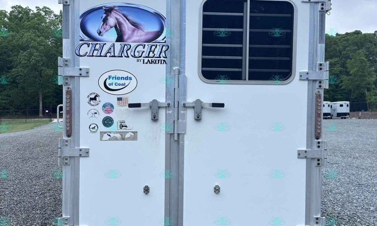 Rear of a white Lakota Charger horse trailer with a large horse logo and various stickers, parked on gravel with trees in the background.