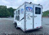 White Lakota Charger horse trailer with horse head logo parked on a gravel lot, with trees in the background.