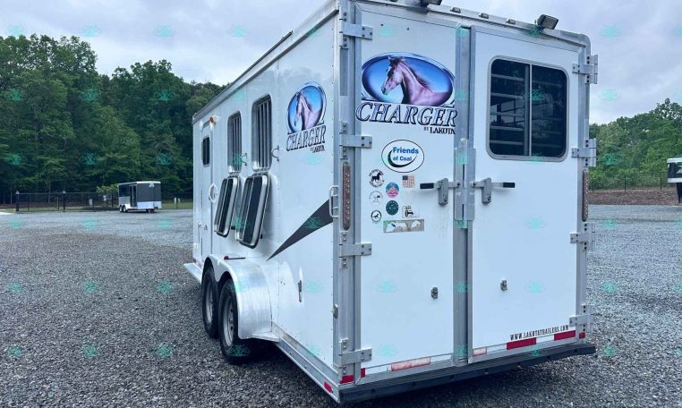 White Lakota Charger horse trailer with horse head logo parked on a gravel lot, with trees in the background.
