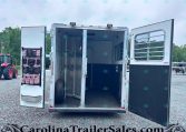 Open horse trailer with left-door tack organizer and empty stalls, parked on a gravel lot outdoors.