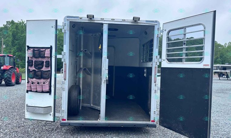 Open horse trailer with left-door tack organizer and empty stalls, parked on a gravel lot outdoors.