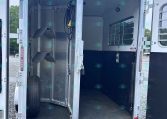 Open aluminum horse trailer interior with stairs on the left, a coiled hose and electrical cord hanging, and a window with bars on the right.
