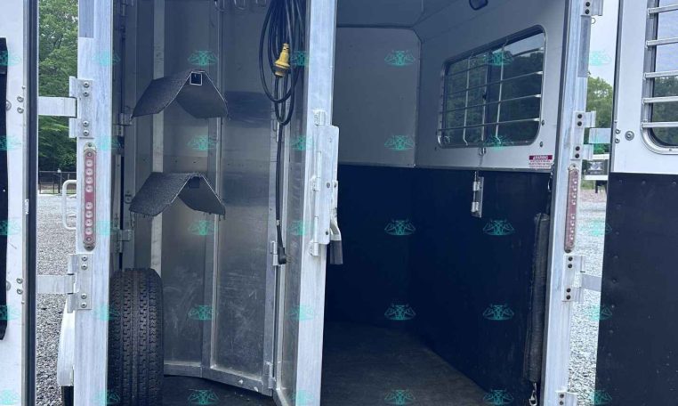 Open aluminum horse trailer interior with stairs on the left, a coiled hose and electrical cord hanging, and a window with bars on the right.