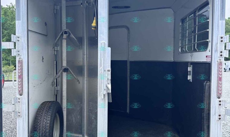 Open horse trailer interior with a center divider, tie rings, and a wheel visible at left, ready for loading.