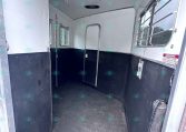 Interior of a horse trailer: white walls, dark lower half, narrow central aisle, and a few small windows. A partition door is visible on the left, and metal bars cover the right window area likely for ventilation and safety.