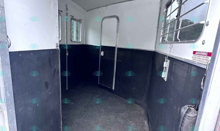 Interior of a horse trailer: white walls, dark lower half, narrow central aisle, and a few small windows. A partition door is visible on the left, and metal bars cover the right window area likely for ventilation and safety.