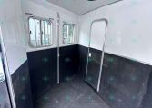 Interior of a small white trailer with two windows and black lower walls, empty stall area.