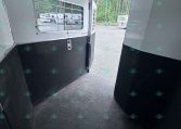 Interior of a horse trailer with white walls and black lower panels, window with bars, and open rear door to a gravel lot with trees and other trailers outside.